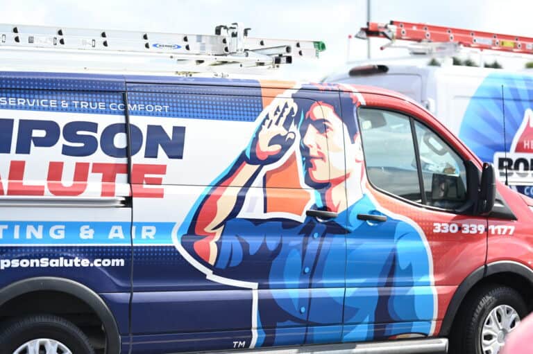 A van with a graphic of a man saluting, advertising a heating and air company. A phone number and part of another service van are visible in the background.