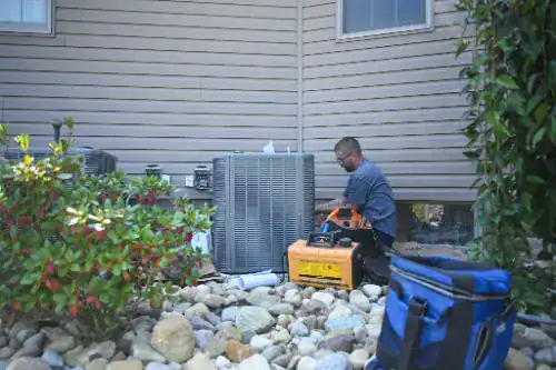 A technician works on an outdoor air conditioning unit beside a house, surrounded by rocks and tools, with plants in the foreground.