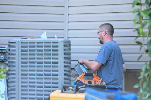 Man in a blue shirt working on an outdoor air conditioning unit next to a house with beige siding.