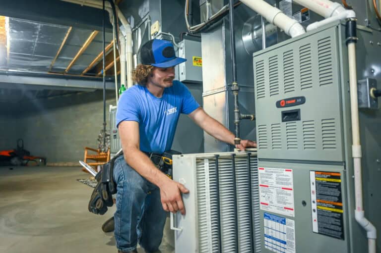 A technician in a blue shirt and cap is servicing a furnace, holding an air filter in a basement utility room.