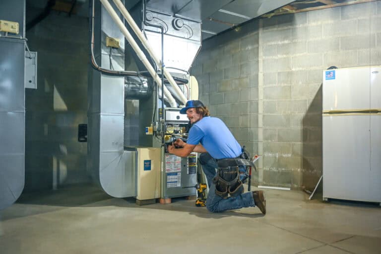 A technician kneels on the floor in a basement, working on a furnace with tools and equipment nearby. Cinder block walls and a refrigerator are visible in the background.