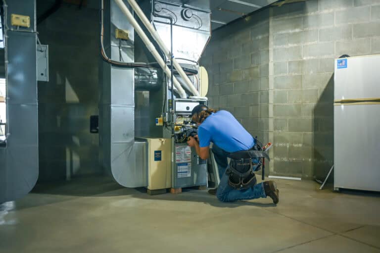 Technician kneeling and inspecting a furnace unit in a basement with concrete walls and floor, next to a refrigerator.
