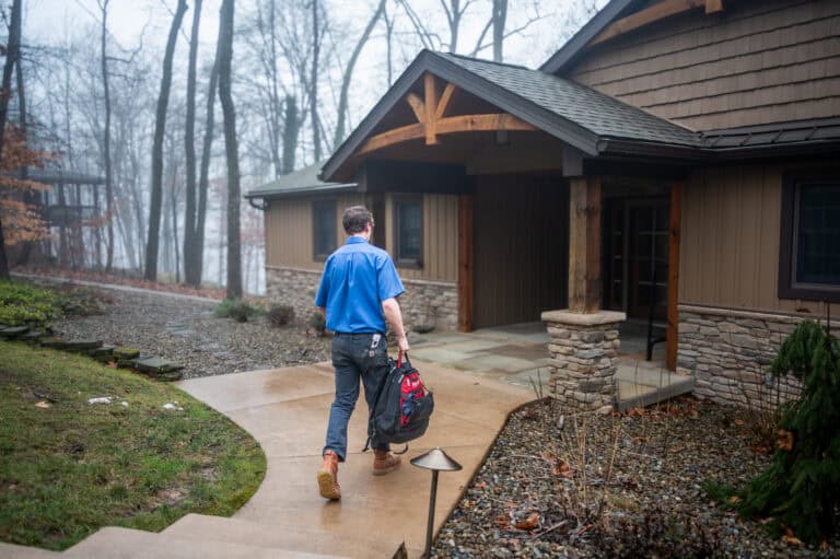 A man in a blue shirt carries a tool bag while walking toward the entrance of a house on a rainy day.